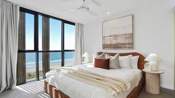 A bedroom with a large bed, two bedside tables, a ceiling fan, and a view of the beach.