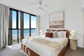 A bedroom with a large bed, two bedside tables, a ceiling fan, and a view of the beach. - Opulent Designer Escape|Ocean View, Pool & Parking (Palm Beach)