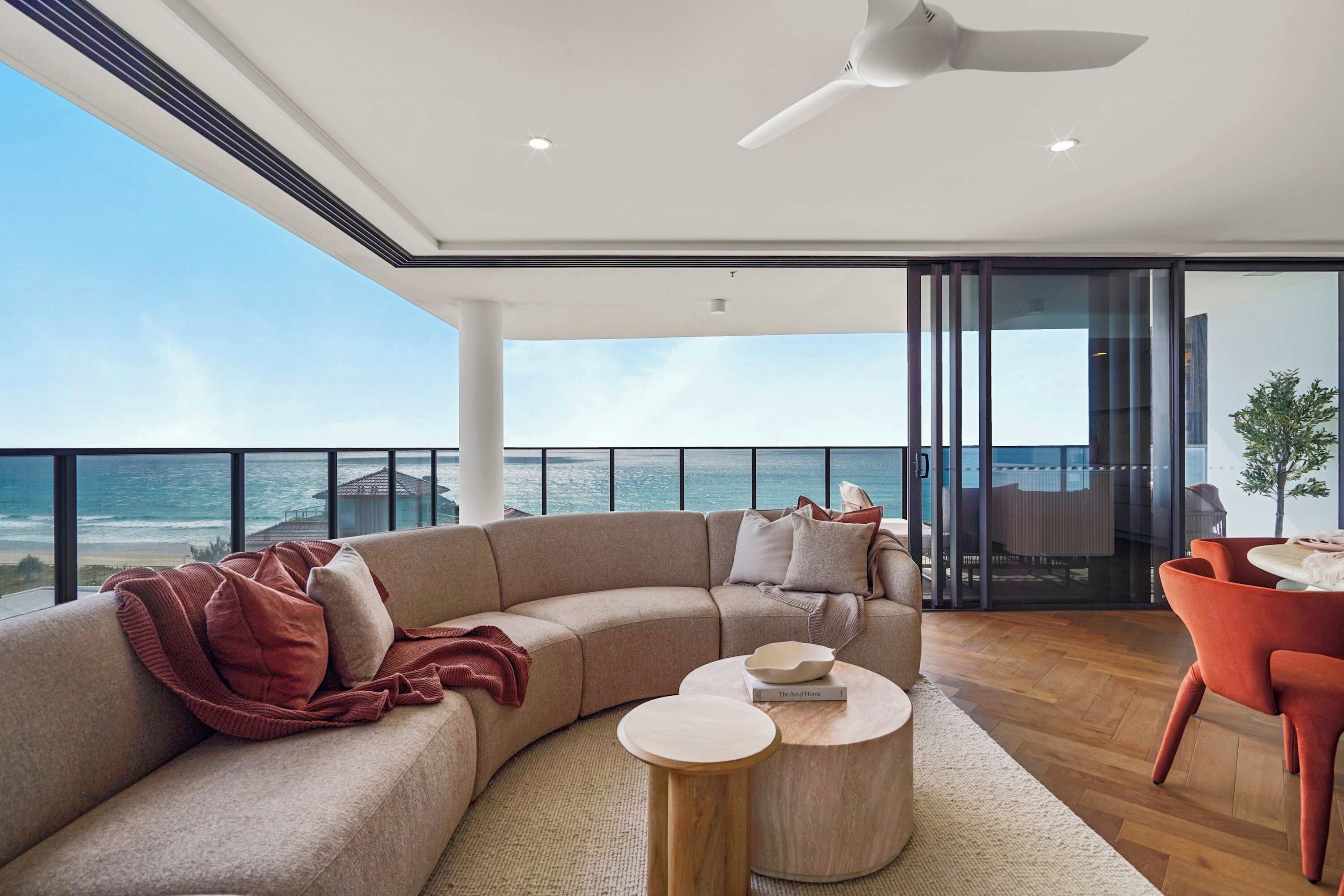 A modern living room with a curved sofa, a round wooden coffee table, and a red chair. A ceiling fan is visible, and the room has large windows offering a view of the ocean.