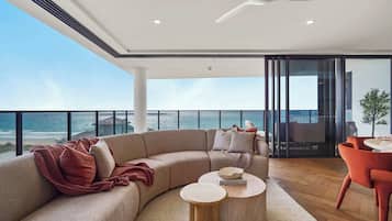 A modern living room with a curved sofa, a round wooden coffee table, and a red chair. A ceiling fan is visible, and the room has large windows offering a view of the ocean.