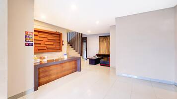 A hotel reception area with a wooden reception desk, a staircase, and a seating area with a sofa and armchairs.