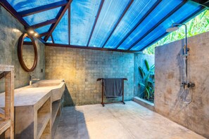 Ein Badezimmer mit einer Betonwand, einem Holzwaschtisch, einem runden Spiegel, einer Dusche mit Glastür und einem Handtuchhalter.