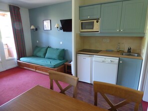 Um espaço compacto para viver, com sofá, mesa de jantar, cozinha equipada com micro-ondas e máquina de lavar louça, e uma janela com cortinas.