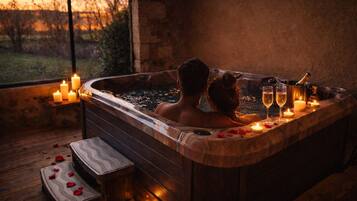 Un couple dans un bain à remous, entouré de bougies et de bouteilles de champagne, avec une terrasse en bois et une vue sur le paysage extérieur.