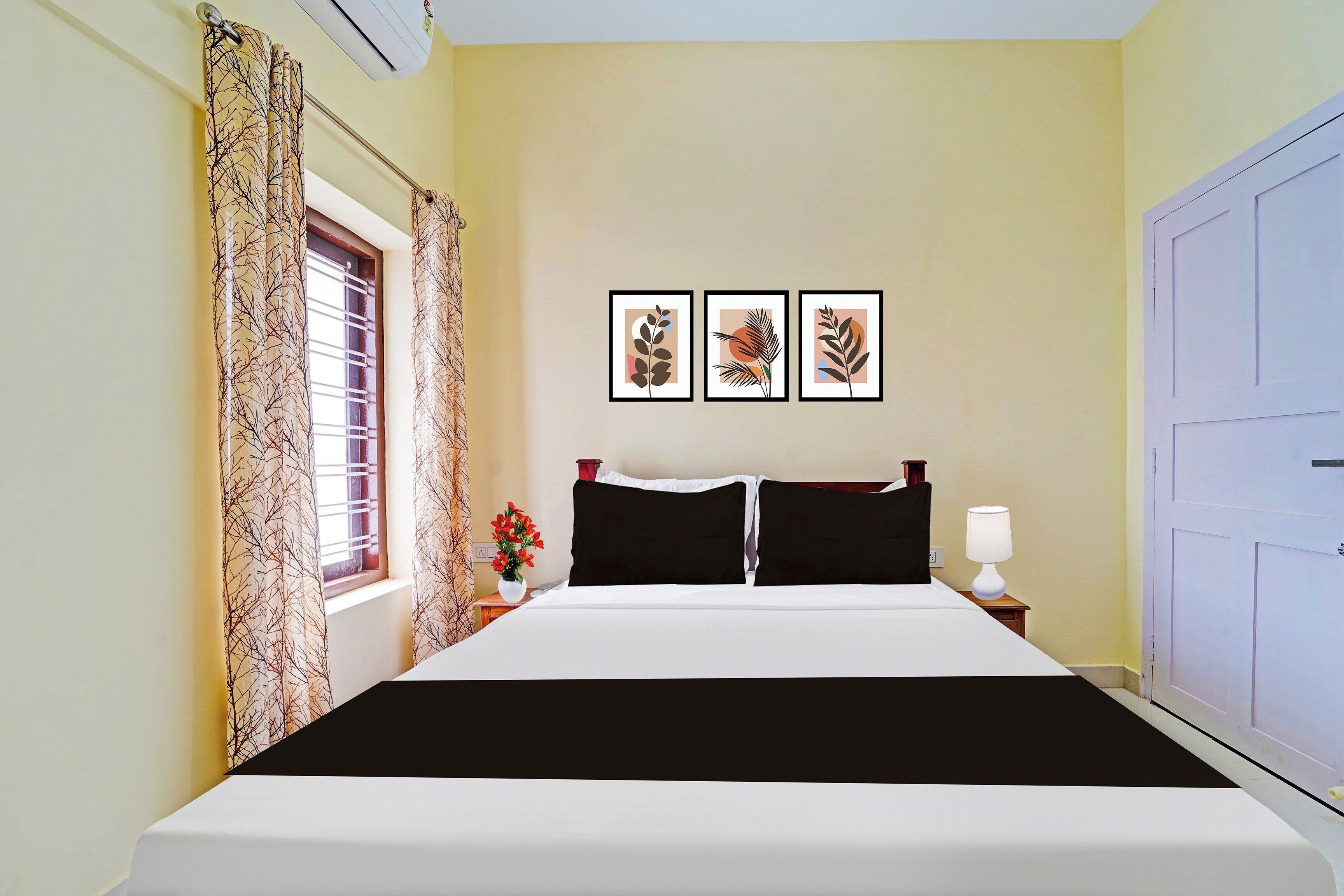 A hotel room with a bed, a nightstand, a lamp, a window with curtains, and three framed pictures on the wall.