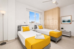 A modern hotel room with two beds, a large wardrobe, a desk, and a window with a view. - 2BR Apartment with Pool - El Breeze by Premier (Pernera)