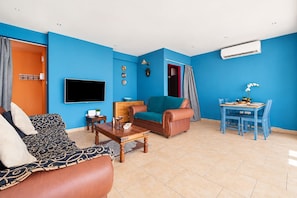 A living room with a sofa, a coffee table, a TV, a dining table with chairs, and a blue wall. - 2BR Apartment with Pool - Aphrodite by Premier (Ayia Napa)