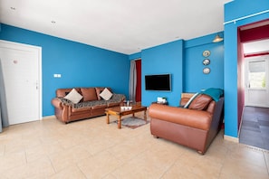 A living room with two leather sofas, a coffee table, a flat-screen TV, and a door with a red curtain. - 2BR Apartment with Pool - Aphrodite by Premier (Ayia Napa)