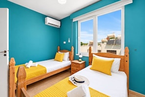 A room with two wooden beds, a window with a view of buildings, and an air conditioning unit. - 2BR Apartment with Pool - Aphrodite by Premier (Ayia Napa)