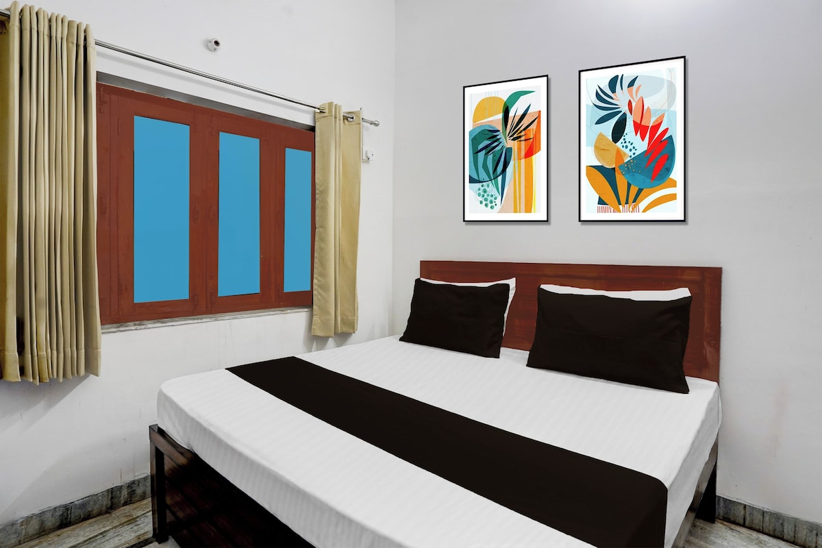Quarto de hotel com cama, duas almofadas, janela com cortinas e duas pinturas abstratas emolduradas na parede.