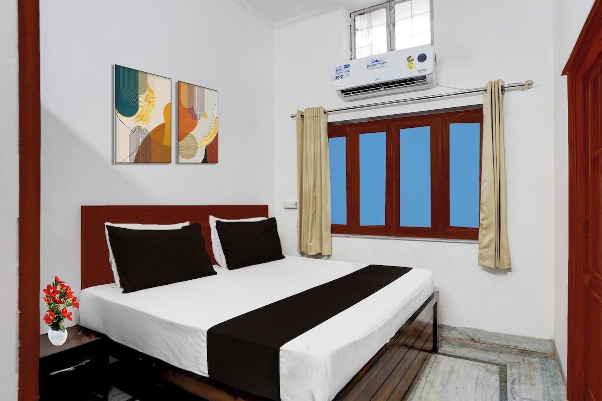 A hotel room with a bed, a window with curtains, an air conditioning unit, and two wall-mounted artworks.