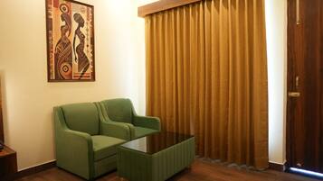 Una habitación con un sofá verde, una mesa de centro de vidrio, piso de madera, cortina marrón y una puerta.