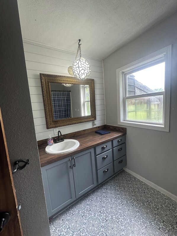 A bathroom with a white sink, gray cabinets, a patterned floor, a large mirror, and a hanging light fixture.