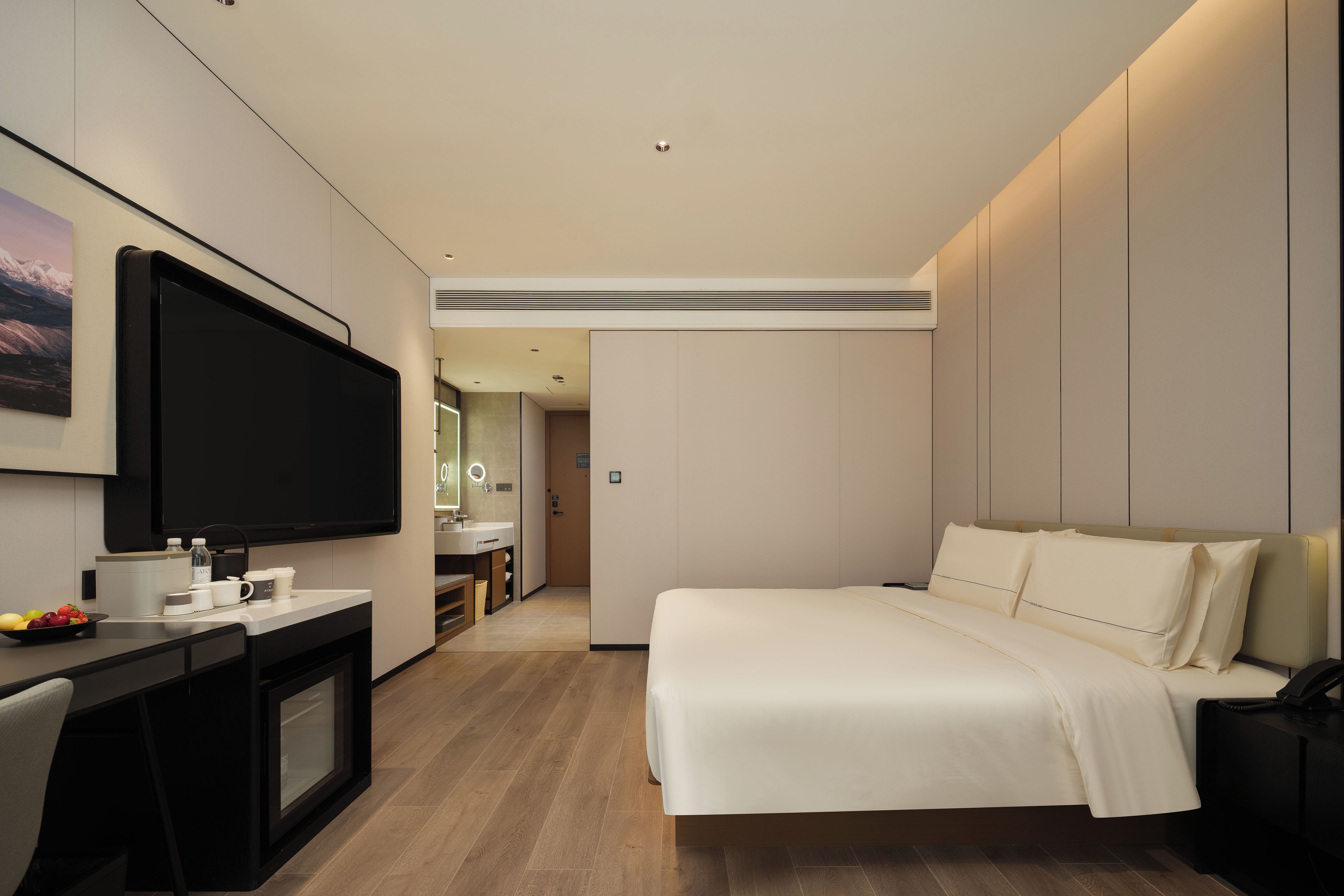 A modern hotel room with a large flat-screen TV, a bed with white linens, a desk with a computer and a cup, and a bathroom visible through an open door.