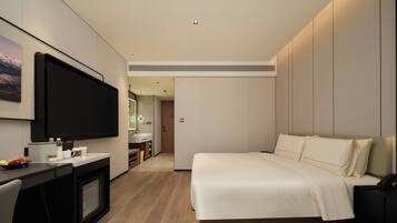 A modern hotel room with a large flat-screen TV, a bed with white linens, a desk with a computer and a cup, and a bathroom visible through an open door.