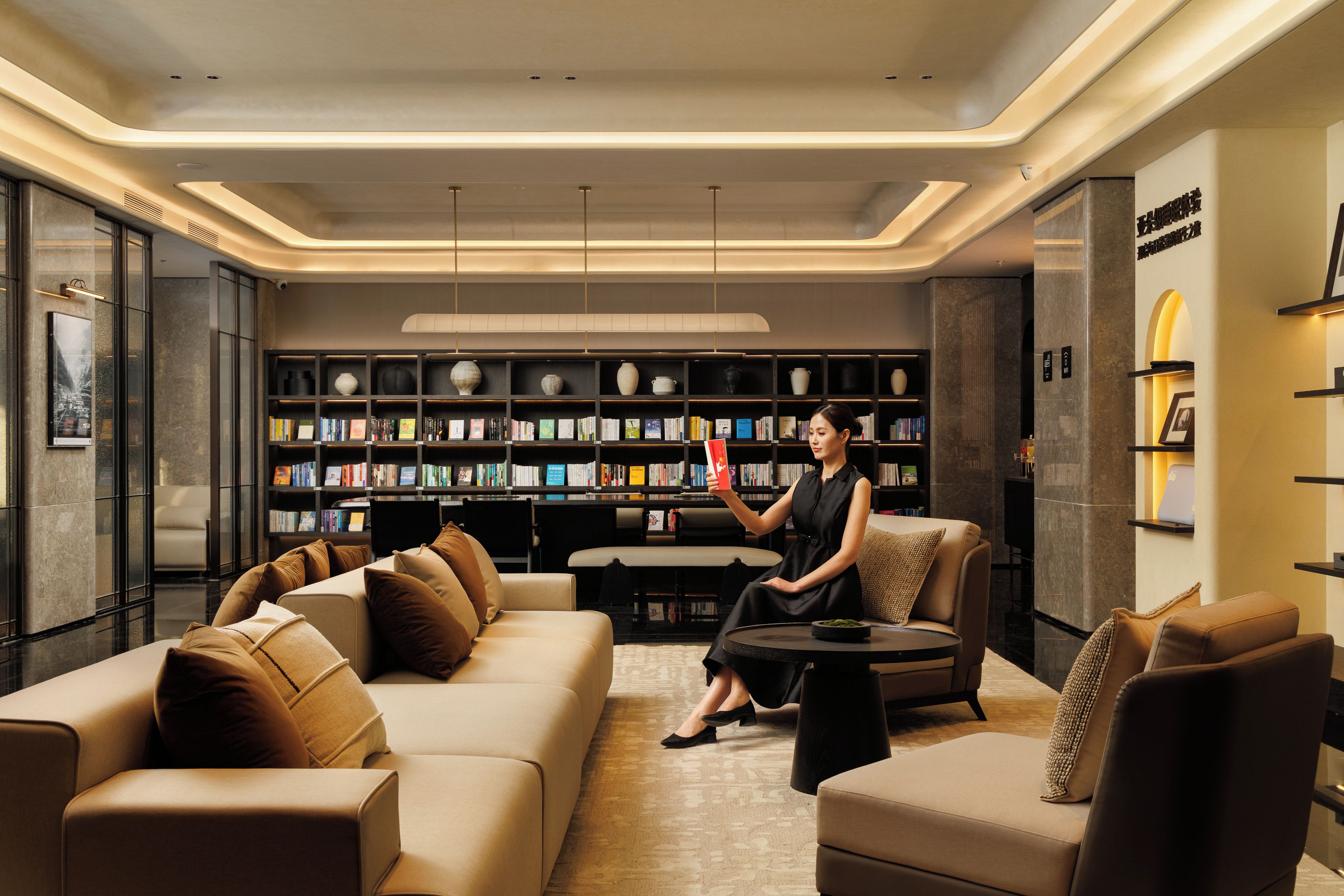 Una biblioteca moderna con sofás de cuero, una mujer leyendo un libro y estanterías llenas de libros y objetos decorativos.