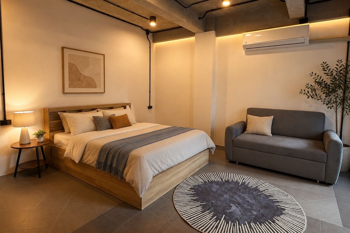 Un dormitorio moderno con una cama, un sofá gris, un tapete redondo y un equipo de aire acondicionado.
