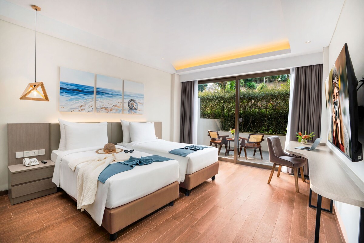 A hotel room with two beds, a desk, a chair, a TV, and a balcony with a view of greenery.