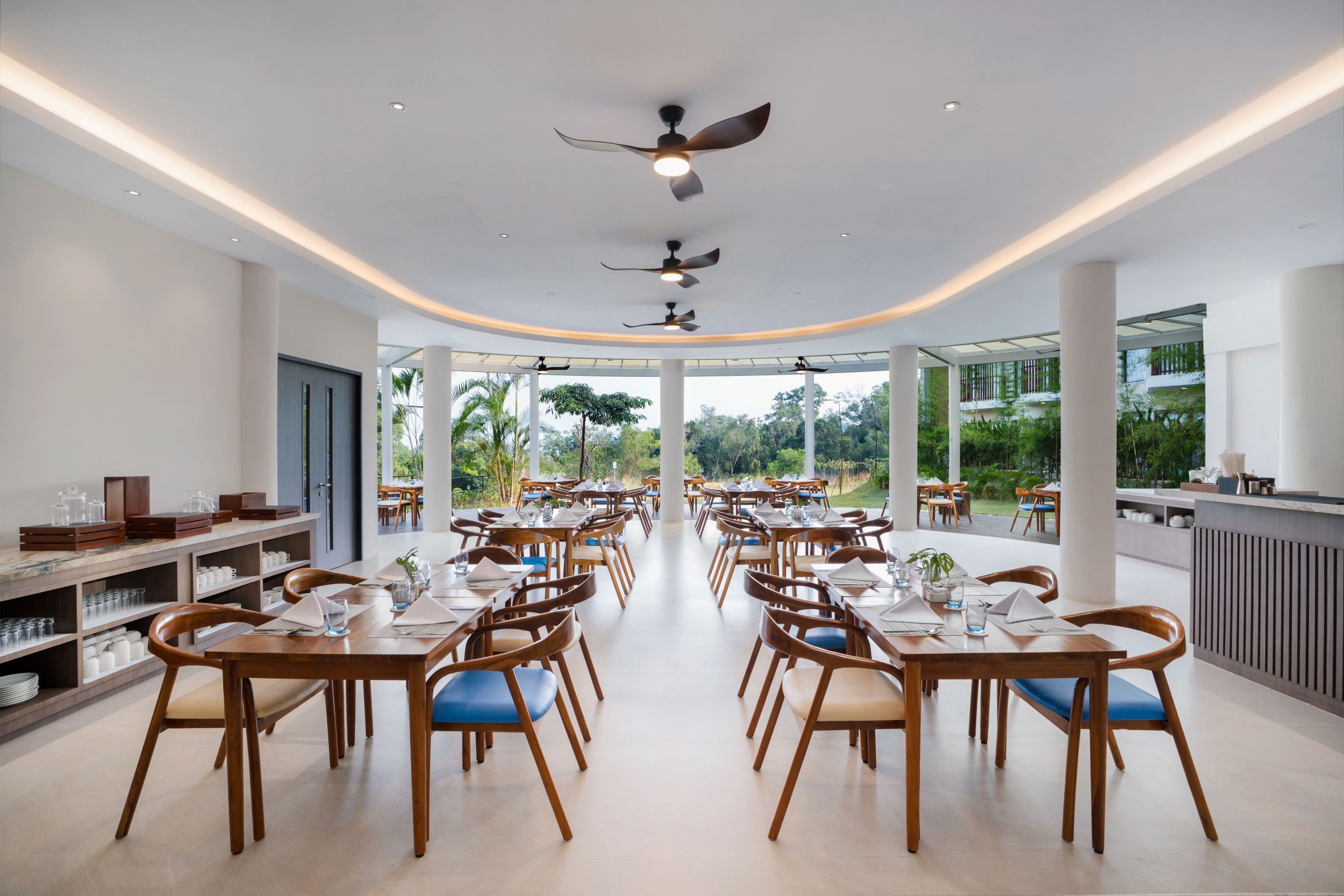 Un vaste espace dédié à la salle à manger, avec plusieurs tables et chaises, des ventilateurs de plafond et une vue sur l’extérieur.