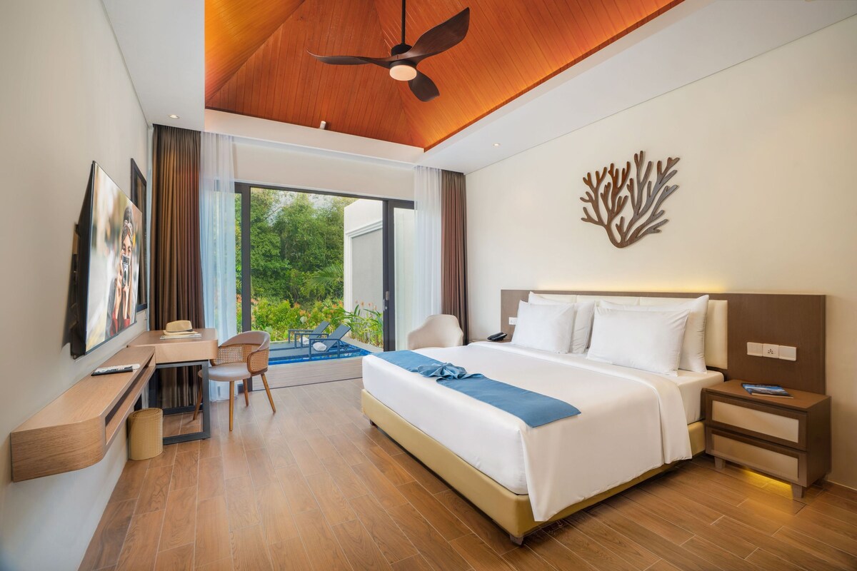 A hotel room with a bed, bedside table, chair, desk, and a view of the outdoors.