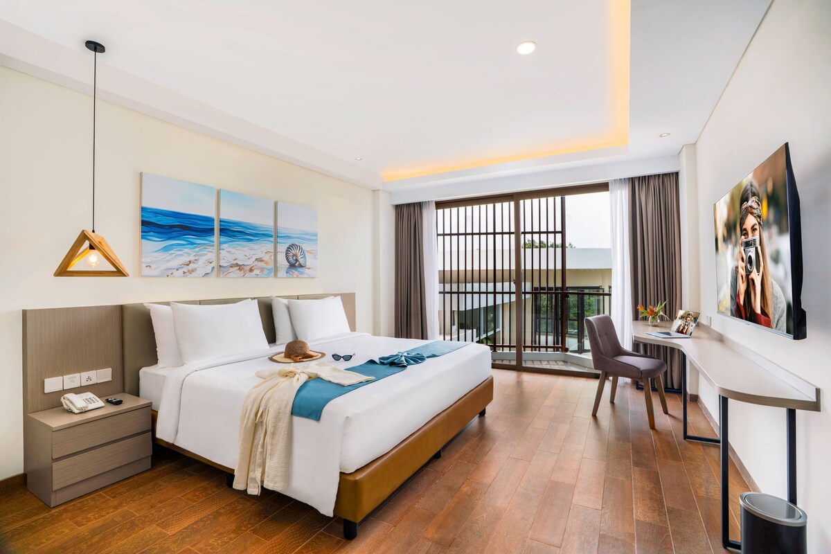 A hotel room with a bed, bedside table, lamp, artwork, and a view of the outdoors.