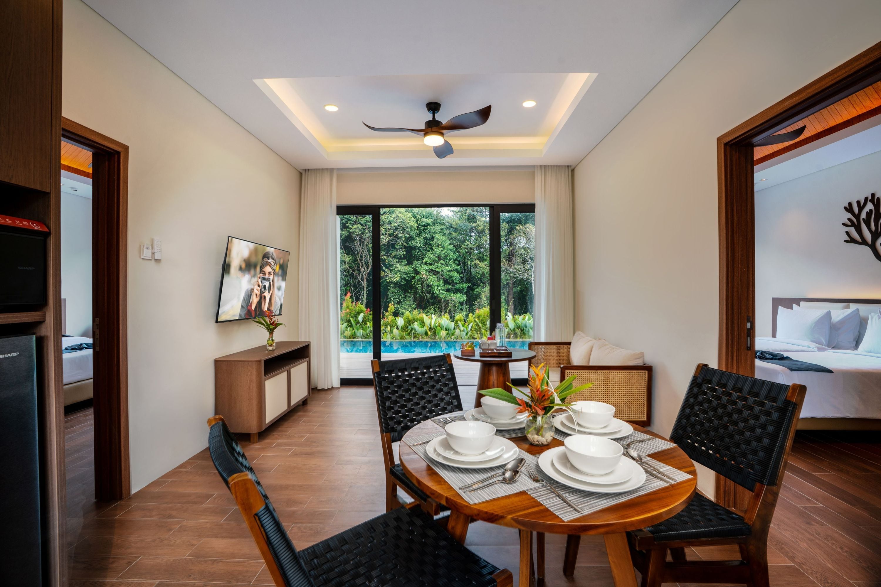 Un coin repas avec une table dressée pour quatre personnes, un ventilateur de plafond et une vue sur une piscine, visible à travers de grandes fenêtres.