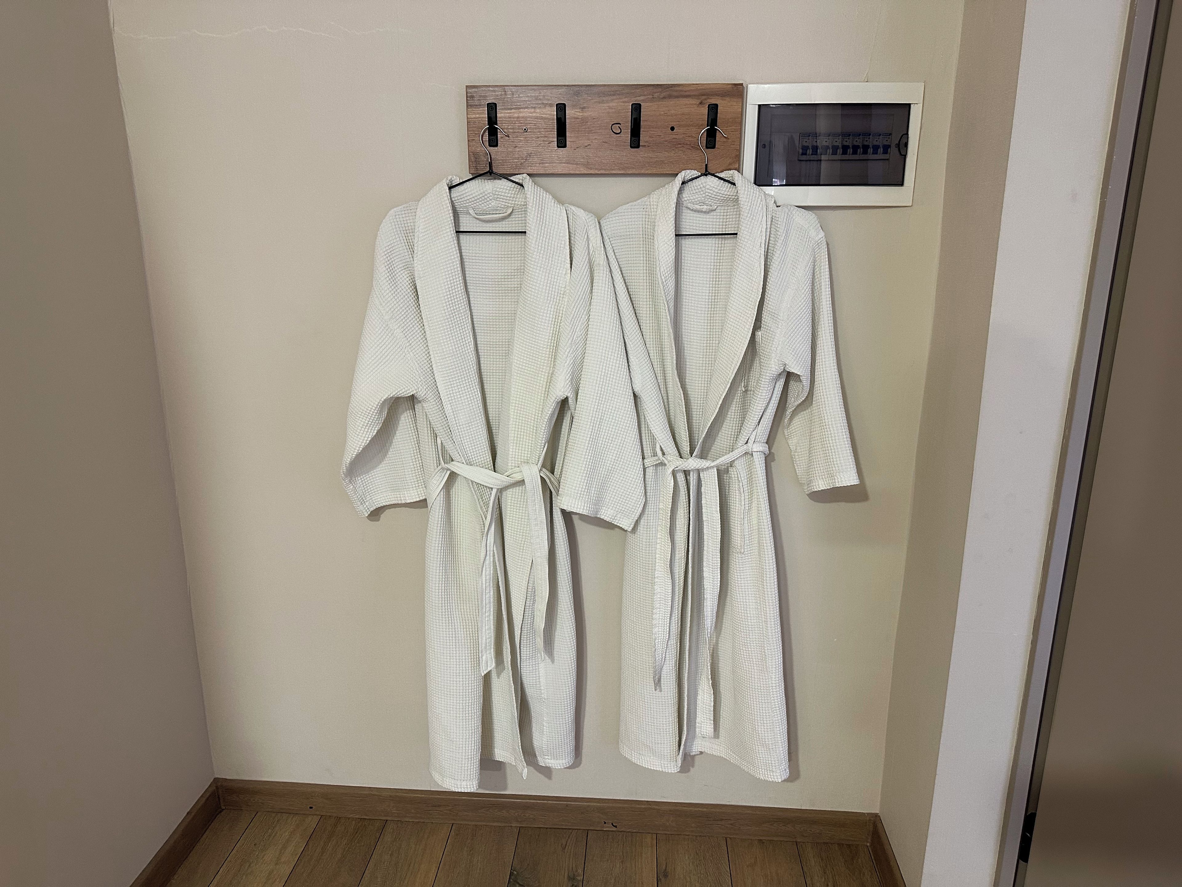 Deux peignoirs blancs sont suspendus à un porte-vêtements en bois, devant un mur et au-dessus d’un plancher en bois.
