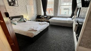 Ein Hotelzimmer mit zwei Betten, einem Schreibtisch, einem Sessel, einem Fernseher und einem Fenster mit Blick auf Gebäude.