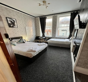 A hotel room with two beds, a desk, a chair, a TV, and a window with a view of buildings. - Standard Triple Room (Eastbourne)