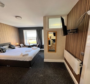 A hotel room with a bed, a TV mounted on the wall, a window with curtains, and a radiator. - Standard Triple Room (Eastbourne)