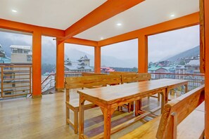 Una habitación con una mesa de madera y bancos, ventanas amplias con vista a edificios y montañas.