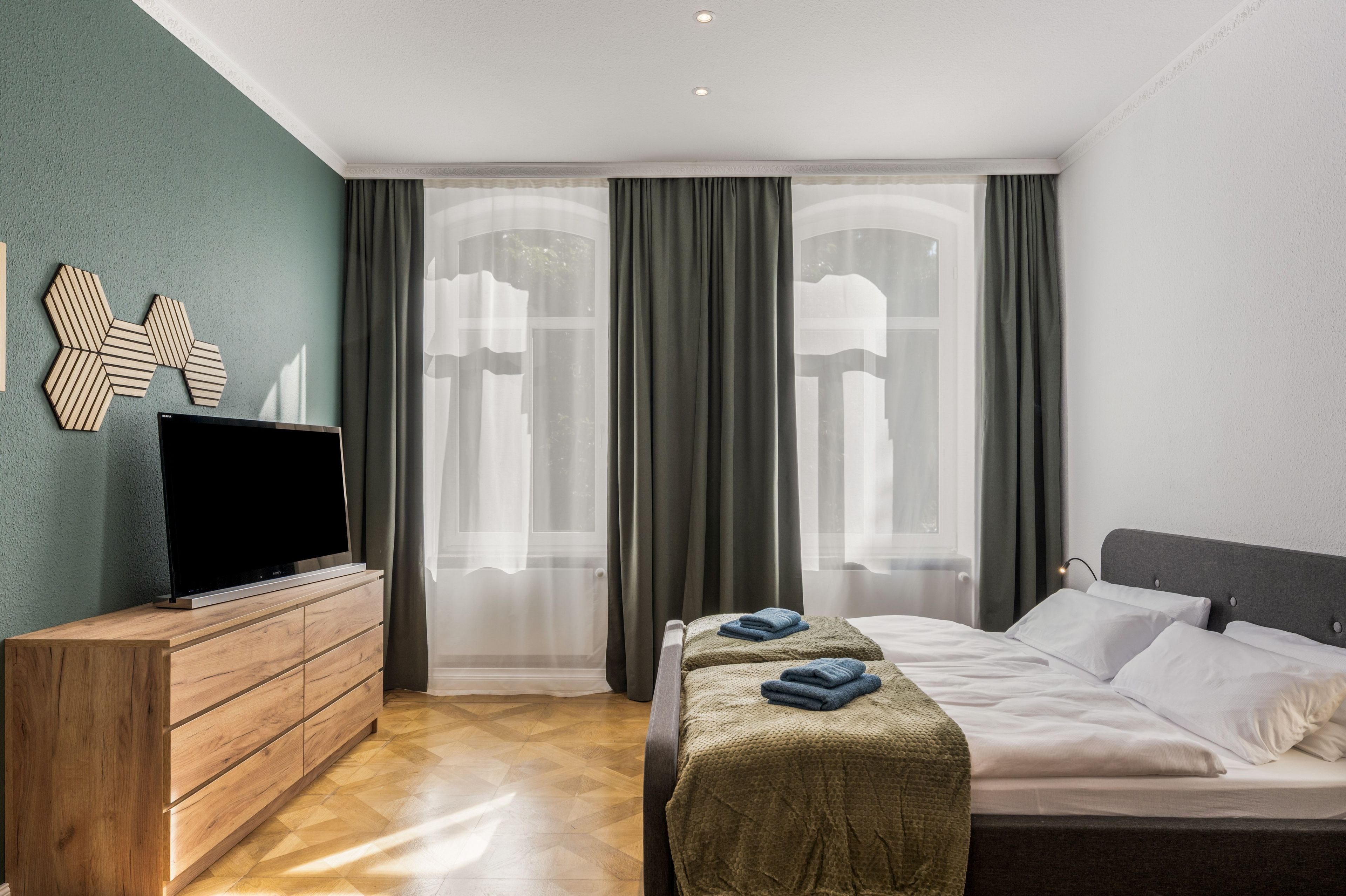Una habitación de hotel con cama, televisor sobre un aparador de madera y ventana con cortinas.