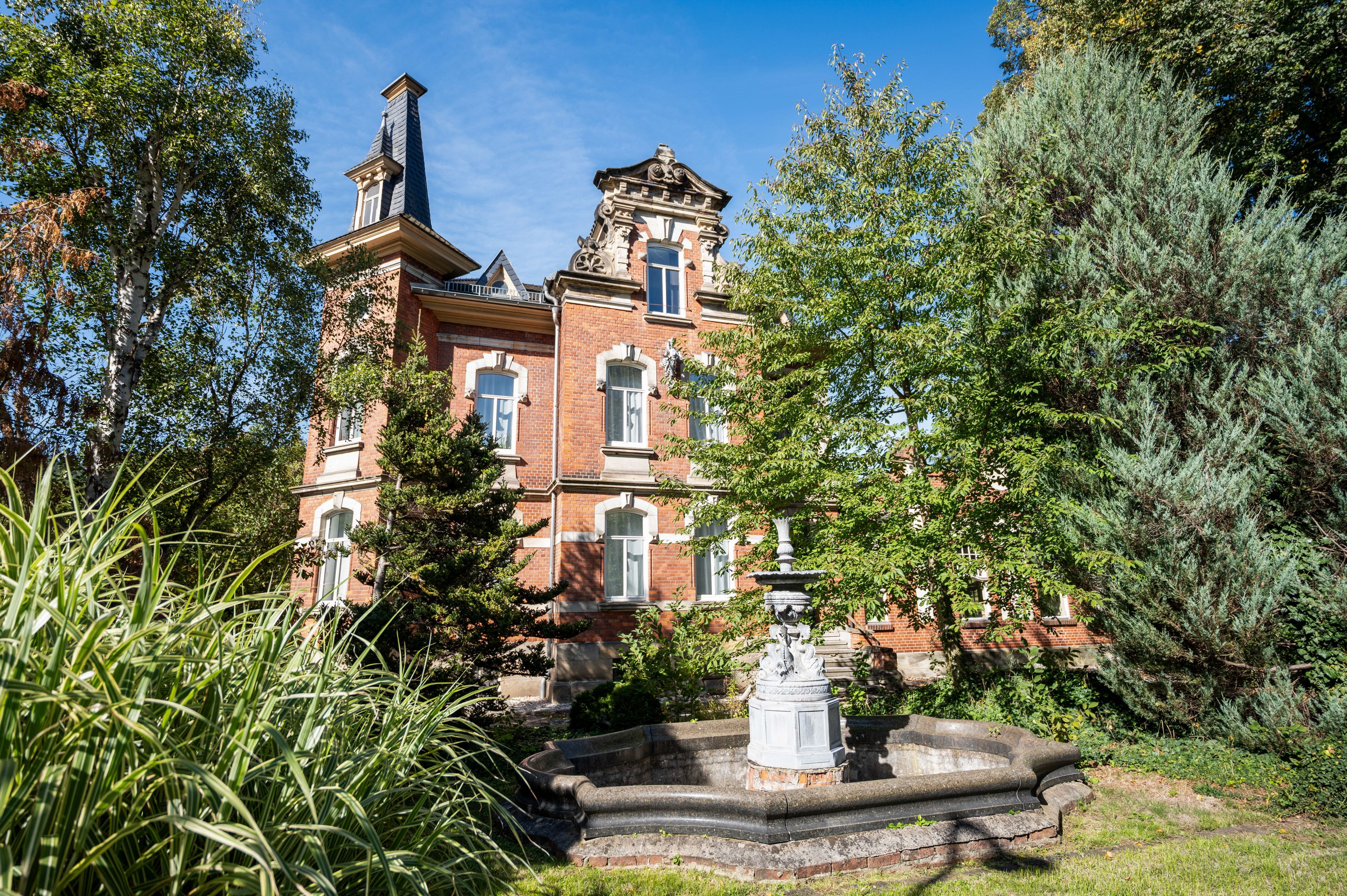 Un grand bâtiment historique, doté d’une tour imposante et de nombreuses fenêtres, entouré d’une végétation luxuriante et agrémenté d’une fontaine en pierre.