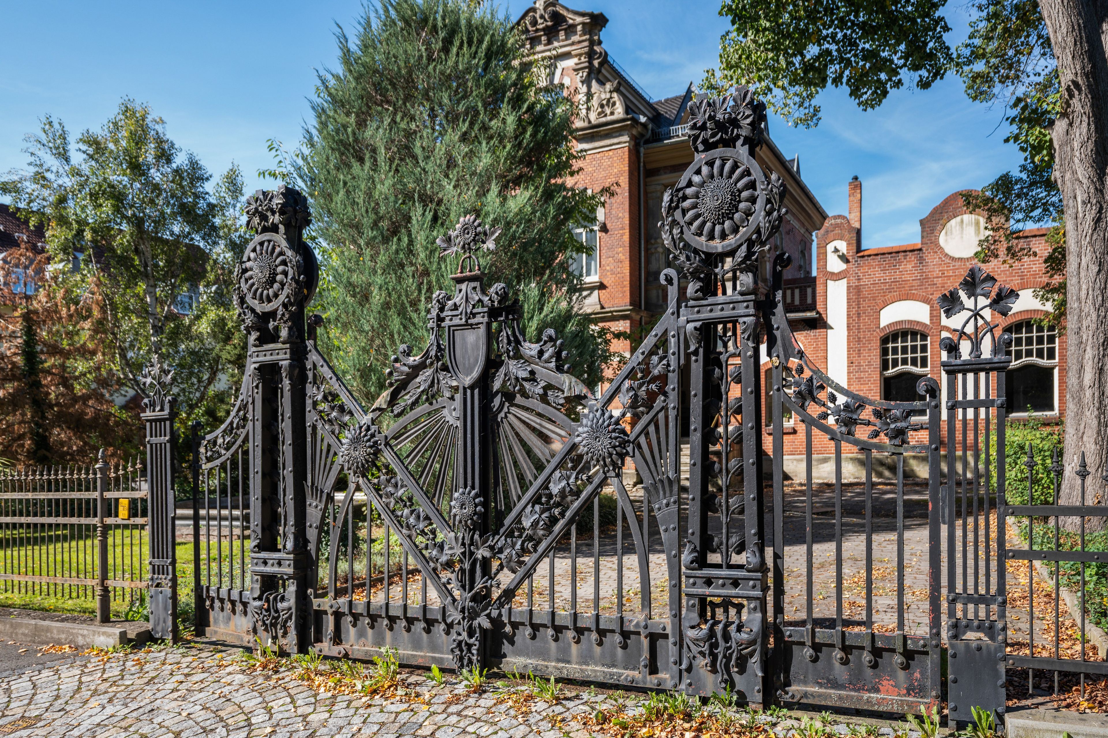 A wrought iron gate with intricate designs, leading to a brick building with a clock tower.