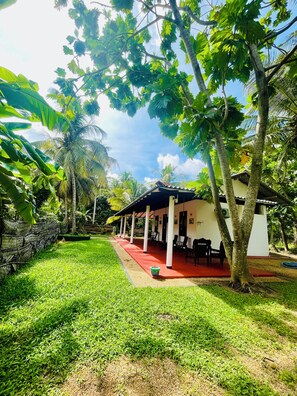 A covered outdoor seating area with a red floor, surrounded by lush greenery and palm trees.
