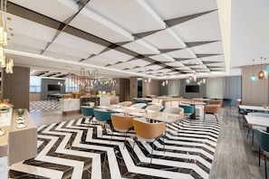 A modern hotel lobby with a geometric ceiling design, marble flooring, and a variety of seating arrangements including tables and chairs.