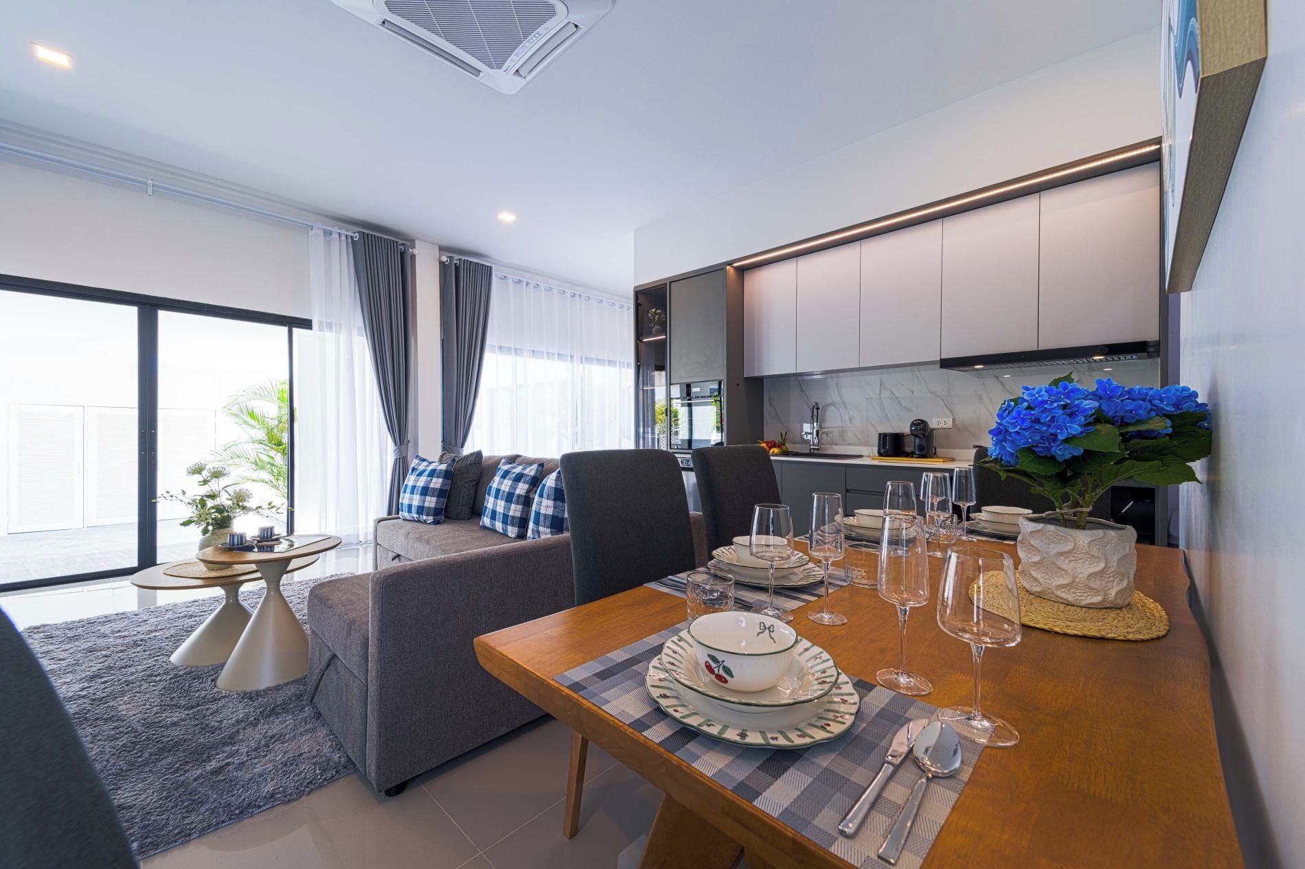 Uma sala de estar moderna com sofá, mesa de jantar posta para quatro pessoas e cozinha ao fundo.