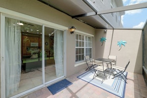 A covered patio area with a sliding glass door leading to a kitchen, a small table with chairs, and decorative palm tree wall art. - Resort Pool, Lanai! Family Retreat Near Disney (Kissimmee)