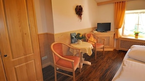 Quarto de hotel com porta de madeira, uma cama, mesa pequena com cadeiras, televisão e janela com cortinas.