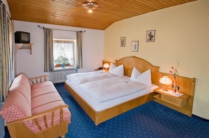 Uma cama de casal com cabeceira de madeira, mesa de cabeceira com abajur, televisão pequena sobre um suporte e uma janela com cortinas.