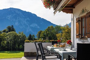 An outdoor seating area with tables and chairs, a mountain view, and a building with wooden shutters. - Double Room No. 14, shower and toilet, balcony, TV, Wi-Fi (Ruhpolding)