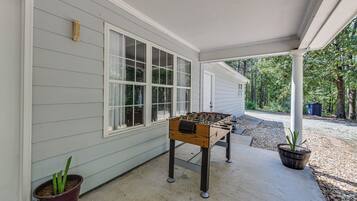 A covered outdoor area with a foosball table, a potted plant, and a house with windows and siding.
