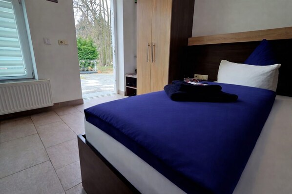A hotel room with a bed, a wooden wardrobe, a radiator, and a view of the outdoors.