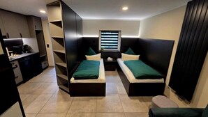 Habitación compacta con dos camas individuales, una kitchenette pequeña y una ventana con persianas.