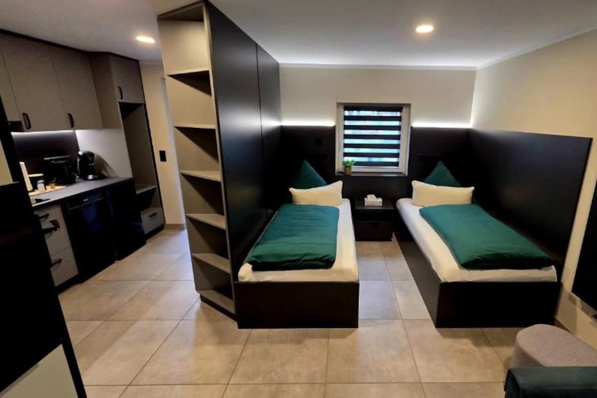 A compact room with two single beds, a small kitchenette, and a window with blinds.