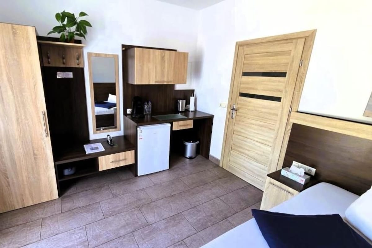 A compact hotel room with a bed, a small kitchenette, a mirror, and a wardrobe.