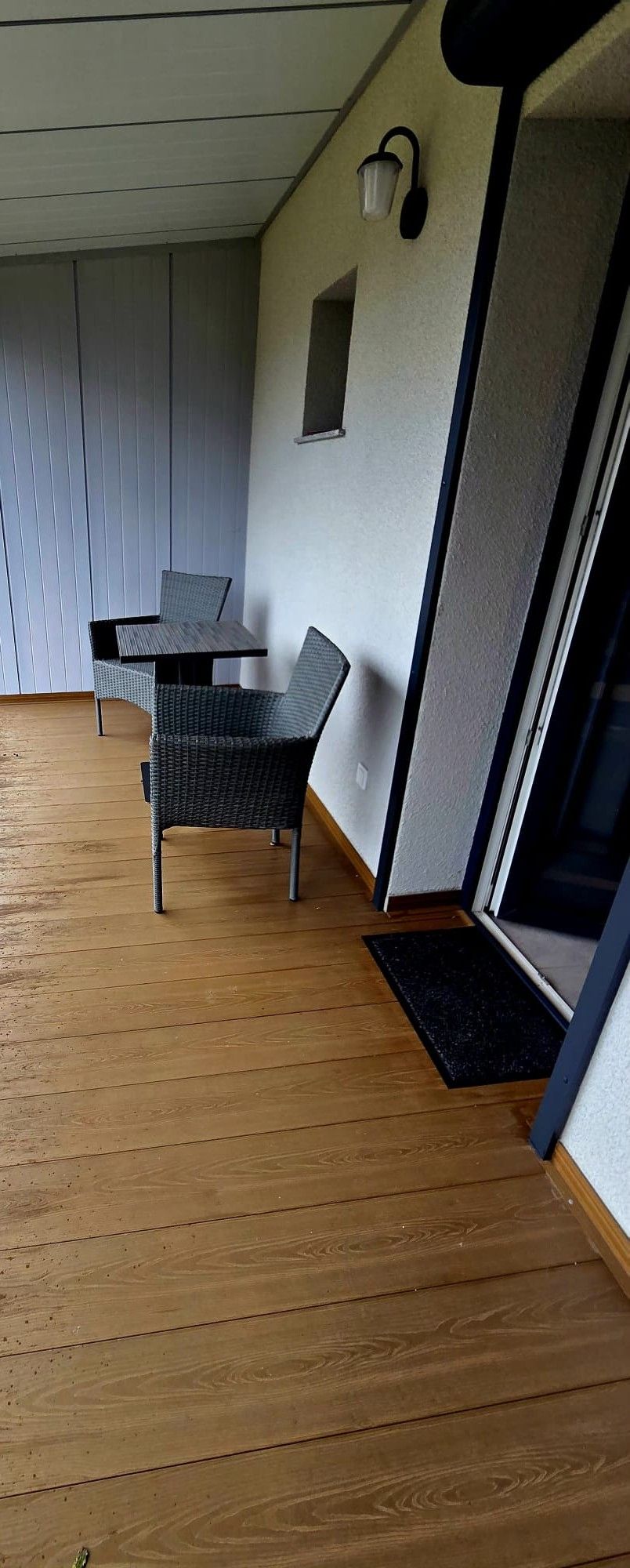 A room with a wooden floor, a table with chairs, a wall-mounted light fixture, and a door with a glass panel.