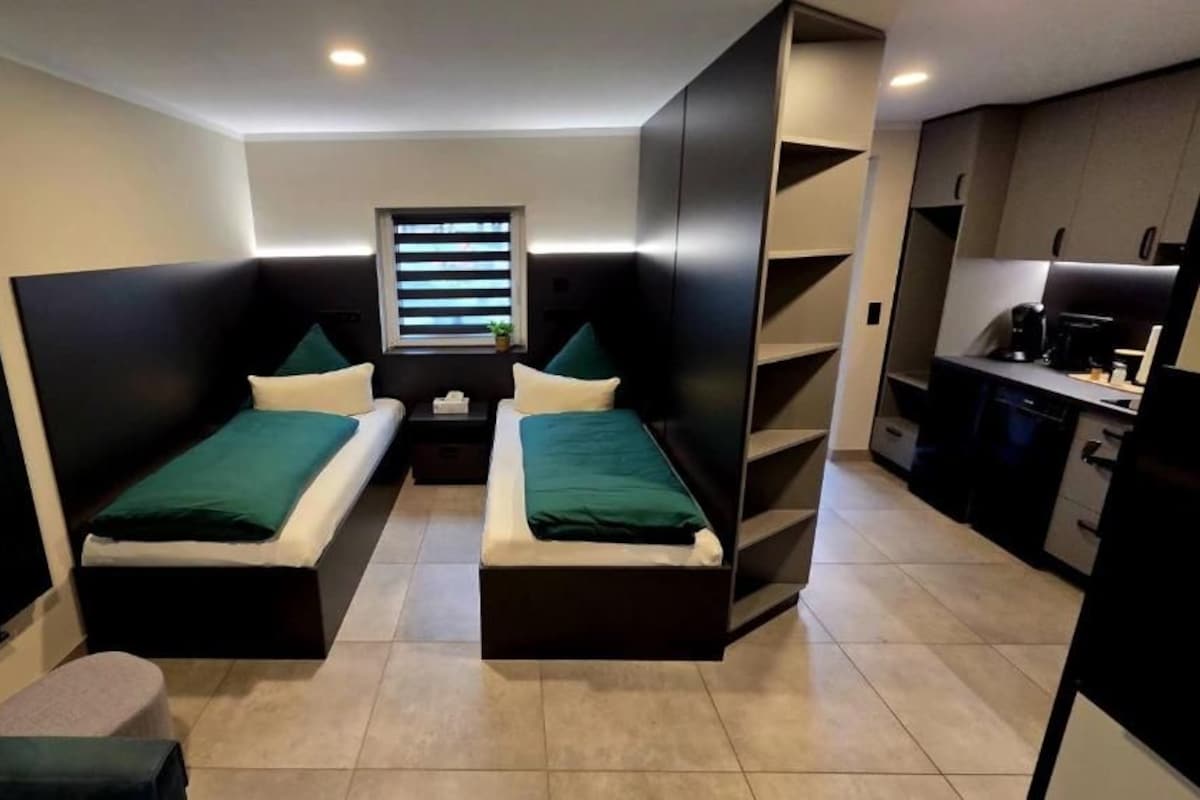 A compact hotel room with two single beds, a small table, a window with blinds, and a kitchenette.