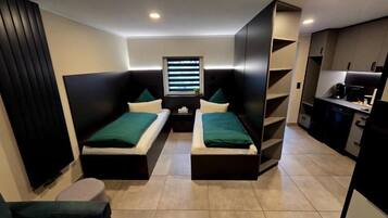 A compact hotel room with two single beds, a small table, a window with blinds, and a kitchenette.