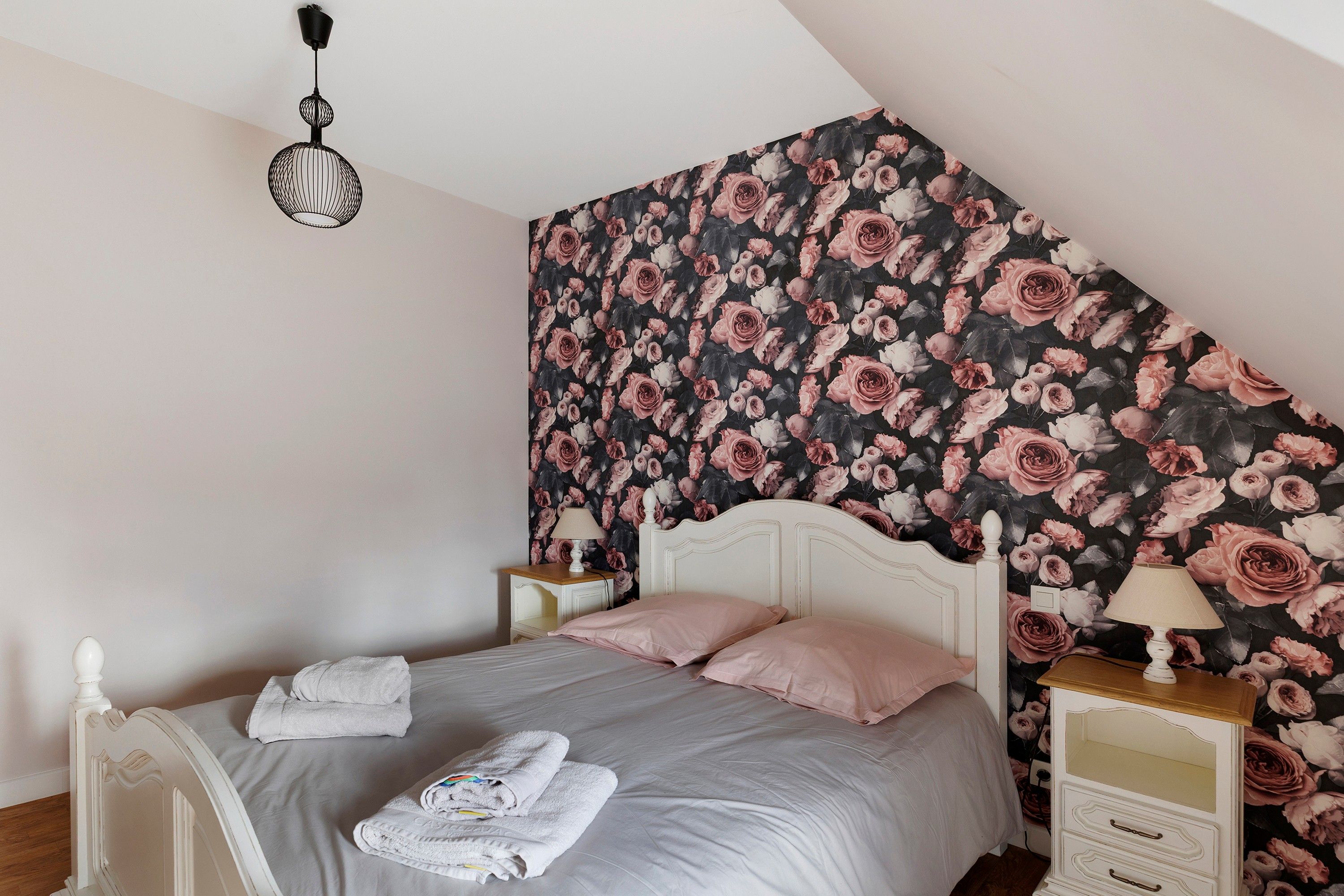 A bedroom with a sloped ceiling, a bed with white bedding, two bedside tables with lamps, and a floral wallpaper.